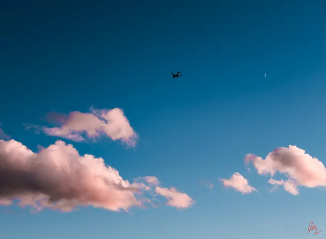 A plane crossing a blue evening sky with pink clouds and a small crescent moon.
